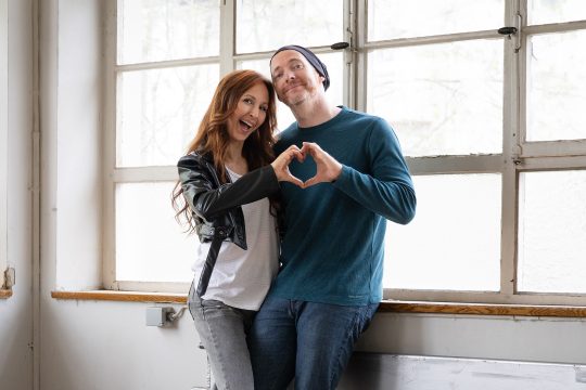Das Duo Perlregen steht vor einer Fensterfront, die beiden bilden mit ihren Händen ein Herz